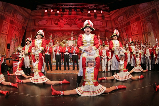 Ein Foto der Tanzgruppe auf der Bühne, im Hintergrund sieht man den stehenden und klatschenden Elferrat, davor stehen Erhengardisten und im Vordergrund stehen die Tanzgruppe und davor die Marieschen im spagat