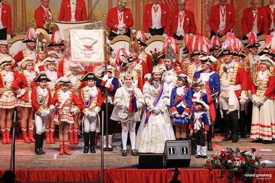 Foto des Cadettencorps mit dem Kinderprinzenpaar auf der Bühne, im Hintergrund auf dem Podest steht der Elferrat, im Vordergrund stehen die Cadetten und das Kinderprinzenpaar