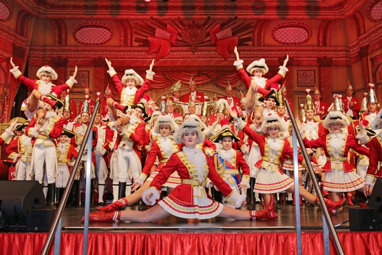 Das Cadettencorps auf der Bühne, im Hintergrund werden Marieschen in einer Hebefigur gehalten und um Vordergrund macht ein Marieschen einen Spagat