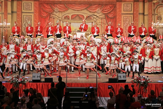 Foto der Bühne der großen Bürgeritzung, im Hintergrund steht der Elferrat auf dem Podest und klatscht während im Vordergrund die Tanzgrupppe alles gibt