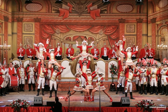 Ein Foto der Tanzgruppe auf der Bühne, im Hintergrund sieht man den stehenden und klatschenden Elferrat, davor stehen Erhengardisten und im Vordergrund stehen die Tänzer der Tanzgruppe mit Hebefiguren