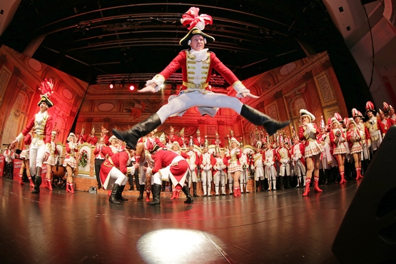 Foto eines Tänzers auf der Bühne, welcher einen beeindruckenden Sprung ausführt, im Hintergrund stehen Klatschende Ehrengardisten