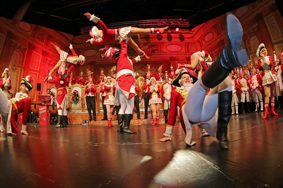 Die Tanzgruppe auf der Bühne beim Tanzen, im Hintergrund stehen die Ehrengardisten und Klatschen, davor werden Marieschen in die Luft geworfen und im Vordergrund tanzen zwei Gardisten