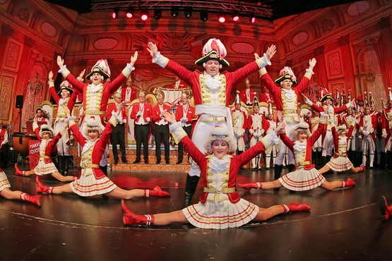Foto der Tanzgruppe auf der Bühne, im Hintergrund steht der Elferrat und im Vordergrund machen die Marieschen einen Spagat