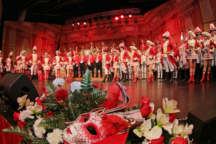 Mitglieder der Ehrengarde Mitglieder der Ehrengarde posieren auf der Bühne. Im Vordergrund sind Blumensträuße sowie eine rot-weiße Maske zu sehen.