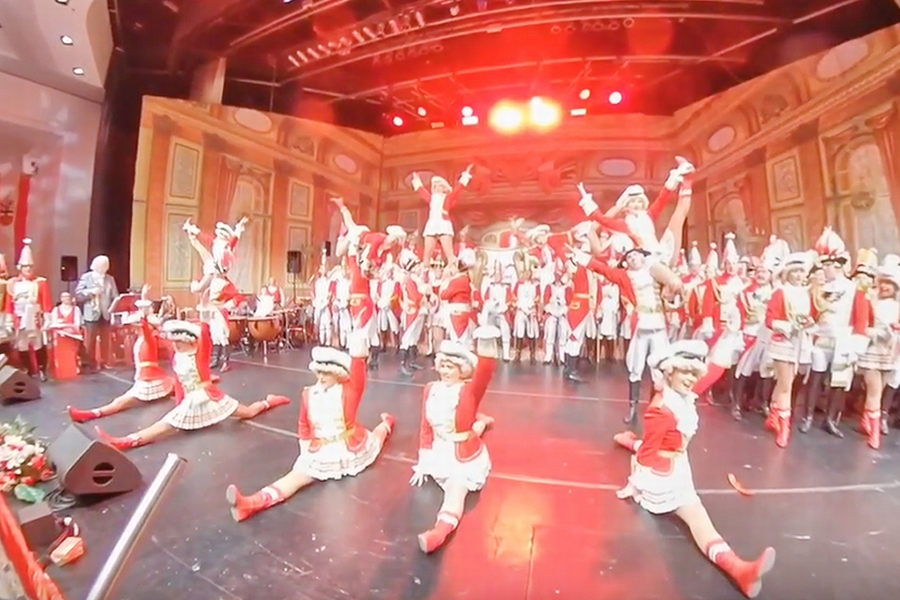 Screenshot aus dem video der Tanzgruppe der EhrenGarden der Stadt Bonn: Im Vordergrund der Bühne führen die Tänzerinnen einen Spagat auf, im Hintergrund heben die Tänzer ihre Tanzpartnerinnen in die Luft